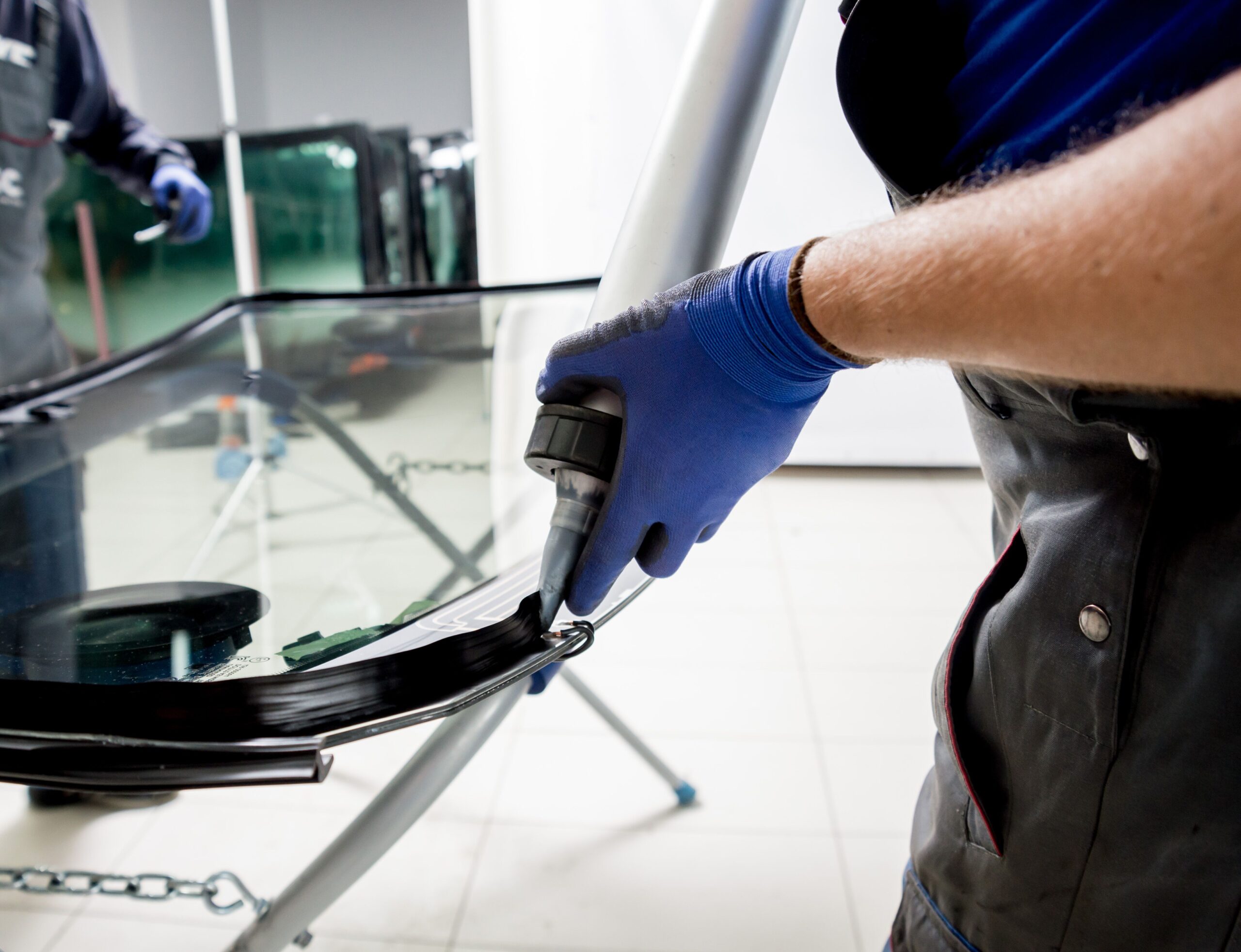 A technician applies adhesive to the edge of a car windshield, preparing it for installation. The setting is a workshop with glass panels in the background.