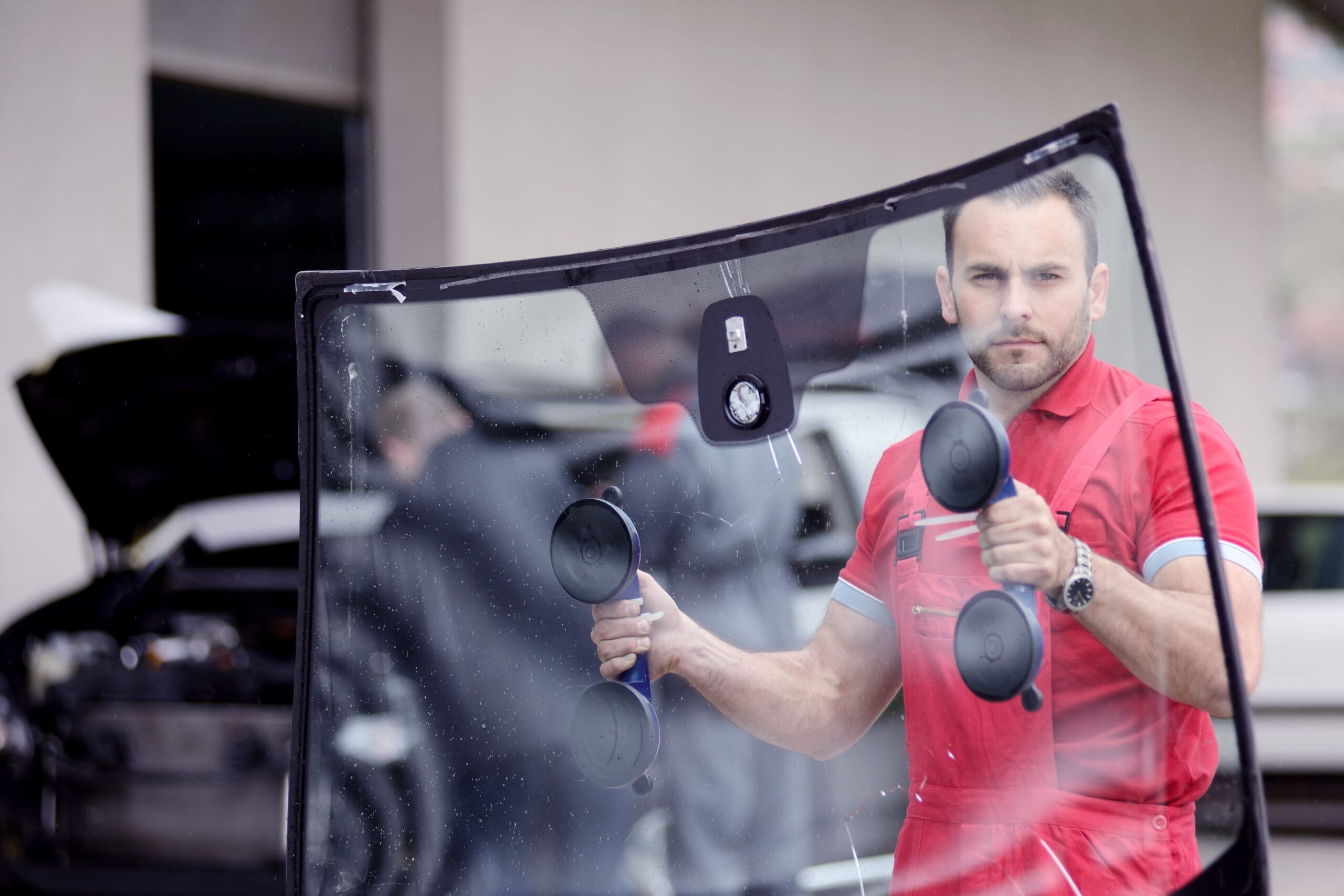 A technician in red overalls holds a large windshield with suction cups, preparing for installation as another worker is visible in the background.