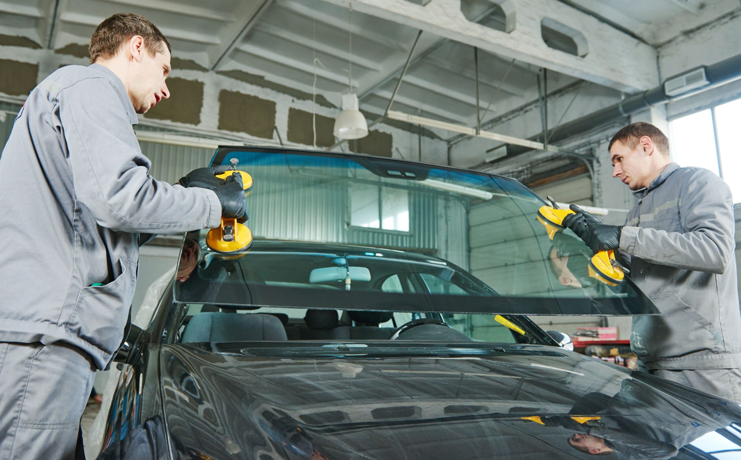 Two technicians replace a car windshield using suction cups in a well-lit garage. Their focus highlights skilled auto glass repair work.